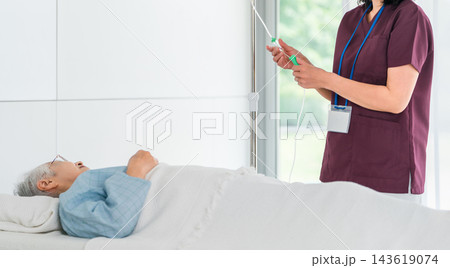 病室でベッドに横たわるシニア女性の点滴を管理するスクラブ姿の看護師 143619074