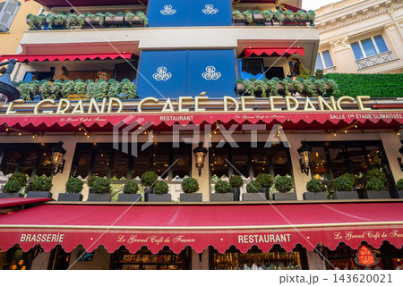 Street level view of Le Grand Cafe de France in Nice, France, with deep red awnings, navy balcony panels, string lights, and layered terraces in early evening. Street level view of Le Grand Cafe de France in Nice, France, with deep red awnings, navy balcony panels, string lights, and layered terraces in early evening. 143620021
