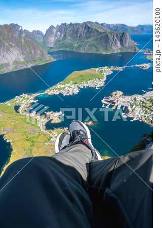 Legs of a person sitting on Reinebringen peak look down upon Reine village, surrounding fjords, and coastal islands in Lofoten, Norway on a clear day. 143620100