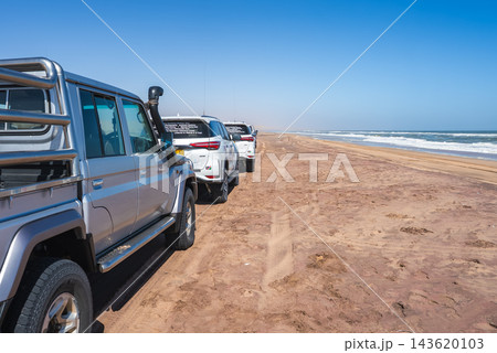 Off road safari vehicles drive and park on the sandy beach along the Atlantic ocean coast in Namibia under a clear sky during daytime. 143620103