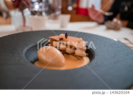 A quenelle of ice cream sits beside a layered tuile with piped cream and petals in a matte black bowl. Warm light and shallow depth of field in Oberlech, Austria. A quenelle of ice cream sits beside a layered tuile with piped cream and petals in a matte black bowl. Warm light and shallow depth of field in Oberlech, Austria. 143620104