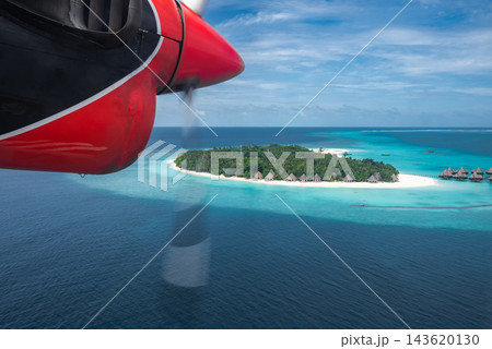Aerial view from a red seaplane engine shows a Maldives atoll with white sand, turquoise lagoons, overwater villas, palm trees, and deep ocean under bright midday light. 143620130