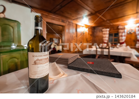 Warm wood stube in Oberlech or Lech, Austria, shows a Schloss Halbturn wine bottle on white tablecloth, thick menus, folded napkin, green tiled Kachelofen, and checkered curtains. 143620184