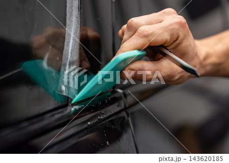 A man carefully applies transparent paint protection film to the exterior black frame of a vehicle door using a turquoise squeegee and precision knife in a car detailing workshop. 143620185