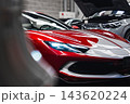 Ferrari 296 GTB sits in a modern workshop, LED headlight on, aero scoop and mesh lower grille visible, SUV with hood up nearby, crisp lighting and shallow depth of field. 143620224