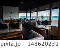 Open air lounge with cream armchairs, dark cushions, and low tables under a high timber roof faces turquoise water, docks, and low islands at a Maldives waterfront. 143620239