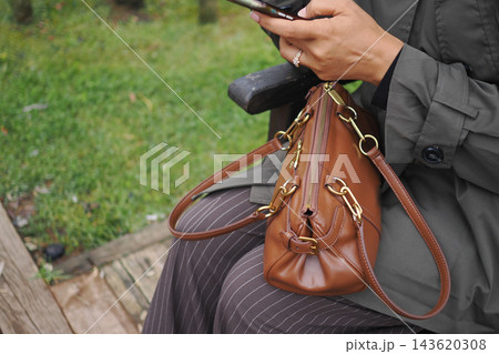 Woman sits outside with a phone and a brown handbag in a green setting Woman sits outside with a phone and a brown handbag in a green setting 143620308