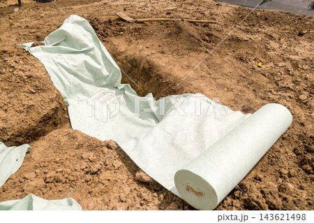 Landscape fabric being carefully placed within a trench filled with rich, dark soil, ready for planting. The outdoor setting is bright and shows the tools used for gardening. Landscape fabric being carefully placed within a trench filled with rich, dark soil, ready for planting. The outdoor setting is bright and shows the tools used for gardening. 143621498