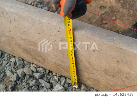 A tape measure stretches out as it is held in a hand, indicating the precise length of a concrete block on a gravel surface at a construction site. A tape measure stretches out as it is held in a hand, indicating the precise length of a concrete block on a gravel surface at a construction site. 143622419