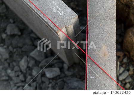 Marking precise alignment with a red chalk line stretched between two concrete blocks on a gravel surface. Marking precise alignment with a red chalk line stretched between two concrete blocks on a gravel surface. 143622420