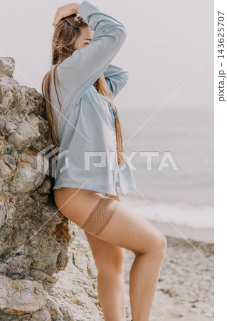Woman beach stockings leaning against a coastal rock while wearing a blue shirt and fishnet hosiery by the ocean Woman beach stockings leaning against a coastal rock while wearing a blue shirt and fishnet hosiery by the ocean 143625707