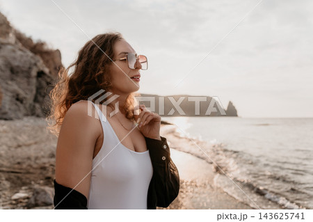 Woman Beach Sunset Woman in sunglasses and white swimsuit stands on a rocky beach at sunset enjoying serene vacation vibes 143625741