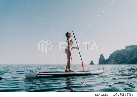Woman Paddleboarding Ocean Coastline: A woman enjoys paddleboarding on a calm ocean, with a stunning coastline in the background. Woman Paddleboarding Ocean Coastline: A woman enjoys paddleboarding on a calm ocean, with a stunning coastline in the background. 143625883