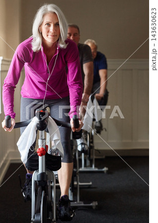 Class, portrait or mature woman on bike machine in workout or training for endurance, health or wellness. Proud, earphones or female sports person on cycling exercise for fitness, challenge or music 143630623