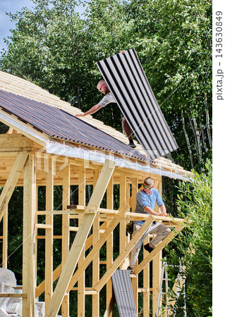 Asphalt roofing sheets are passed to worker on sloped timber roof while another builder handles panels during installation of roofing. 143630849