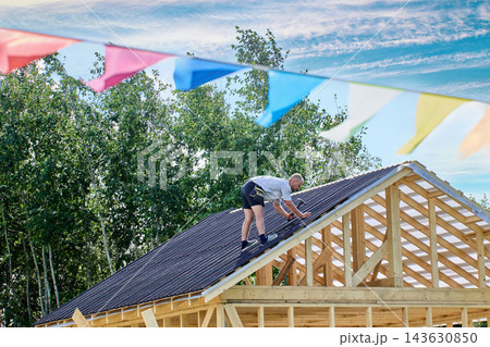 Corrugated bitumen sheets are fixed by worker using nails and hammer on roof frame during construction of wooden building. Corrugated bitumen sheets are fixed by worker using nails and hammer on roof frame during construction of wooden building. 143630850