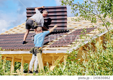 Bitumen corrugated sheets covering part of pitched roof while two men lift another panel into place over exposed wooden battens. Bitumen corrugated sheets covering part of pitched roof while two men lift another panel into place over exposed wooden battens. 143630862