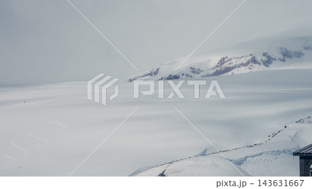 Vast snowfield with distant skiers and glacier-covered mountain slope under overcast sky 143631667