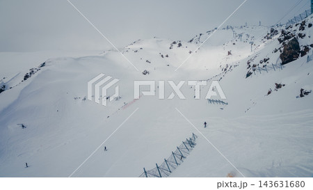 Mount Elbrus snowy ski slope with scattered skiers, rocky ridges, safety fences and chairlift towers under overcast sky 143631680