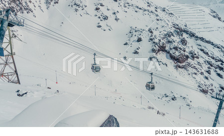 Two gondola cable cars crossing snowy Mount Elbrus slope with rocky ridges and skiers below 143631688