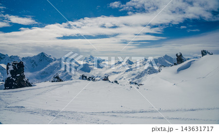 Panoramic snow-covered summit ridge with distant jagged peaks under blue sky and clouds Panoramic snow-covered summit ridge with distant jagged peaks under blue sky and clouds 143631717