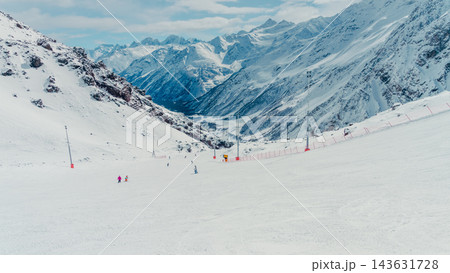 Wide snowy ski slope with skiers and lift poles framed by towering snow-covered Caucasus mountain peaks in crisp daylight 143631728