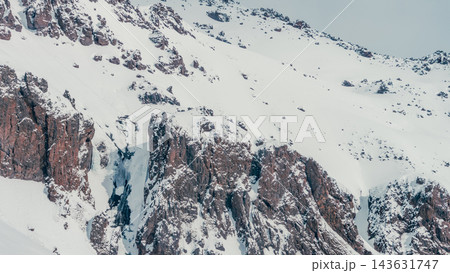 Snow-covered rocky slopes and steep cliffs of high mountain massif under pale winter sky 143631747