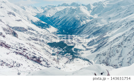 Panoramic snowy alpine valley with ski resort village nestled between towering snow-covered peaks in bright winter daylight and mo 143631754