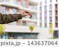 Female Homeowner Holding Keys With House Keychain Against Blurred Apartment Building. New Home Purchase, Mortgage And Relocation Concept. Copy Space. 143637064