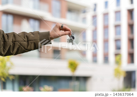 Female Homeowner Holding Keys With House Keychain Against Blurred Apartment Building. New Home Purchase, Mortgage And Relocation Concept. Copy Space. 143637064