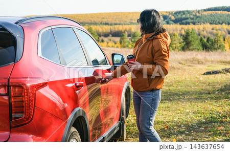 Woman opening red car door holding coffee cup during autumn road trip. Traveler stopped in countryside with forest landscape background 143637654