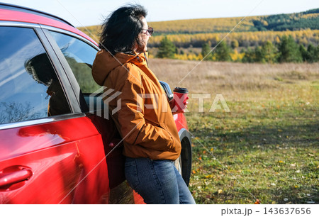 Woman standing near red car holding coffee cup during autumn road trip, resting and enjoying scenic autumn landscape. Travel and freedom concept 143637656