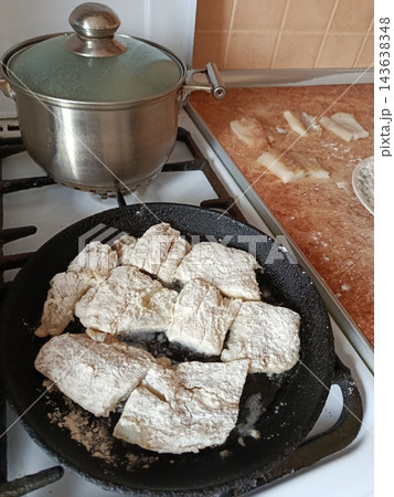 Fish fillets coated in flour cooking in a hot cast iron frying pan on a gas stove burner next to a lidded pot, preparing a delicious seafood meal Fish fillets coated in flour cooking in a hot cast iron frying pan on a gas stove burner next to a lidded pot, preparing a delicious seafood meal 143638348