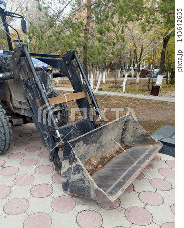 Detailed close-up of a blue and black front loader with its bucket resting on circular paving stones in an urban park. Hydraulic pistons and background trees are visible. 143642635