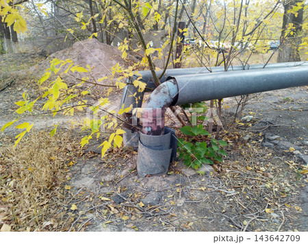 Weathered metal utility pipes with damaged insulation elbows run along the ground in a neglected, autumnal outdoor setting near trees. Weathered metal utility pipes with damaged insulation elbows run along the ground in a neglected, autumnal outdoor setting near trees. 143642709