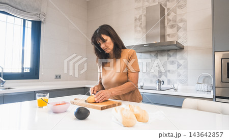 Woman cutting bread loaf as she prepares breakfast at kitchen island Woman cutting bread loaf as she prepares breakfast at kitchen island 143642857