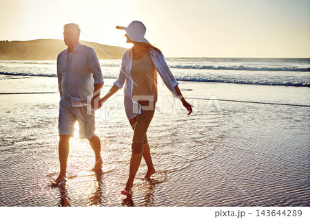 Senior couple, holding hands and walking with beach sunset for bonding, love or support together in nature. Man, woman or lovers enjoying stroll on outdoor summer holiday by water, sea or ocean coast 143644289