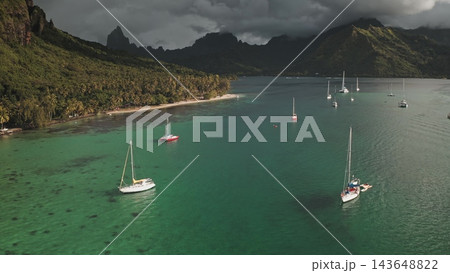 Many sailboats moored in clear turquoise Opunohu Bay, Moorea, French Polynesia, with a palm-fringed sandy beach and lush green mountains beneath a dramatic cloudy sky. Aerial view drone flight 143648822