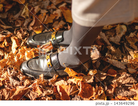 Black loafers with gold chain buckle, woman walking on autumn leaves. Stylish footwear detail, gray tights, fallen foliage on ground. Fashion close up, trendy metallic accessory, fall mood. Black loafers with gold chain buckle, woman walking on autumn leaves. Stylish footwear detail, gray tights, fallen foliage on ground. Fashion close up, trendy metallic accessory, fall mood. 143649296