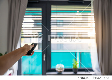 Hand holds a remote control to operate motorized window blinds in a modern apartment. Smart home automation allows convenient adjustment of daylight, privacy and indoor comfort 143649528