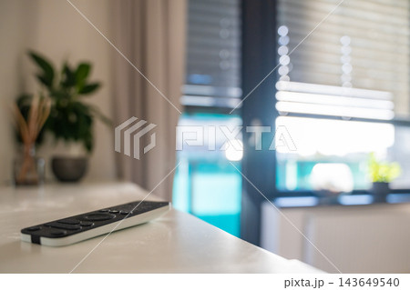 Remote control for motorized window blinds lies on a shelf with blurred blinds in the background. Smart home automation allows convenient daylight control and sun protection with selective focus Remote control for motorized window blinds lies on a shelf with blurred blinds in the background. Smart home automation allows convenient daylight control and sun protection with selective focus 143649540