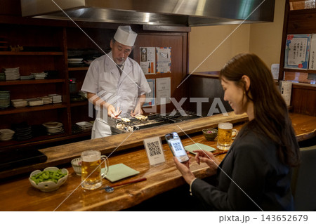 スマホでQRコードを読み込むカウンターの女性客の前で焼き鳥を焼く店主 スマホでQRコードを読み込むカウンターの女性客の前で焼き鳥を焼く店主 143652679