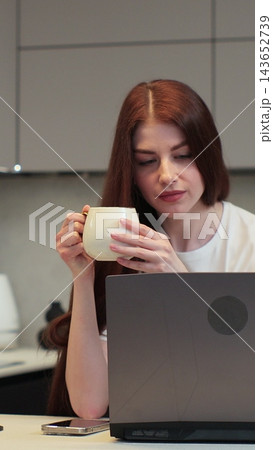 Vertical video. Young woman using laptop computer to browse internet while drinking coffee and working from home in kitchen. Vertical video. Young woman using laptop computer to browse internet while drinking coffee and working from home in kitchen. 143652739