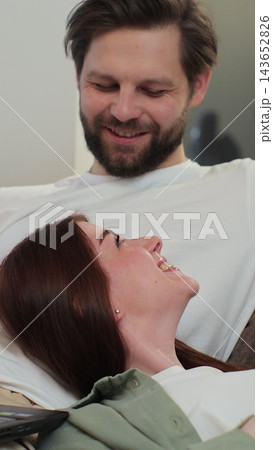 Vertical video. Couple relaxing on sofa, man working on laptop, woman kneeling on man, scrolling social media on mobile phone at home. Concept of remote. 143652826