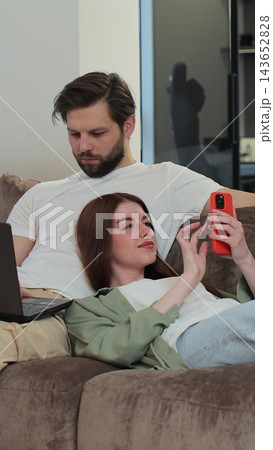 Vertical video. Happy family couple relaxing on sofa, man working on laptop, woman, wife, kneeling on man, scrolling social media on mobile phone at home. Vertical video. Happy family couple relaxing on sofa, man working on laptop, woman, wife, kneeling on man, scrolling social media on mobile phone at home. 143652828
