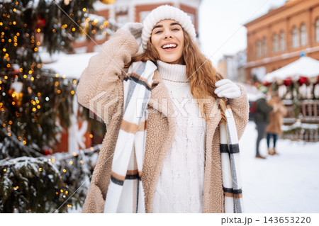Smiling woman in a scarf and hat is having fun at a street holiday fair. Christmas concept. Smiling woman in a scarf and hat is having fun at a street holiday fair. Christmas concept. 143653220
