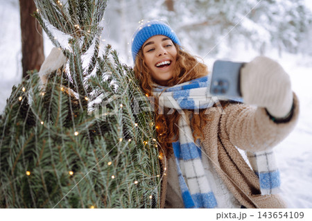 A young woman with a phone and a Christmas tree with garlands enjoys the beautiful winter weather. A young woman with a phone and a Christmas tree with garlands enjoys the beautiful winter weather. 143654109