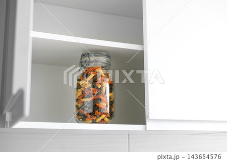 Glass jar filled with dry tricolor fusilli pasta inside kitchen cupboard 143654576