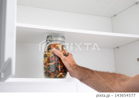 Hand taking glass jar of tricolor fusilli pasta from kitchen cupboard 143654577