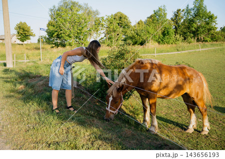 Young woman petting a grazing horse beside a fenced pasture on a sunny rural farm. Horse farm visits rural tourism and animal care Young woman petting a grazing horse beside a fenced pasture on a sunny rural farm. Horse farm visits rural tourism and animal care 143656193